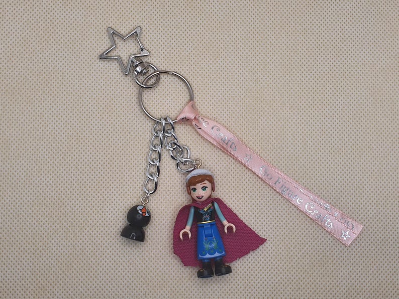 Puede incluir: Un llavero plateado con un cierre en forma de estrella, una cadena y una peque&ntilde;a figura negra. Incluye una figura en miniatura de Anna de la pel&iacute;cula Frozen, con un vestido azul y una capa rosa. Se adjunta una cinta rosa con texto.