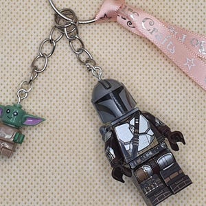 Puede incluir: Un llavero con dos figuras estilo Lego: El Mandaloriano y Grogu. La figura del Mandaloriano viste un traje plateado y marr&oacute;n. Grogu es verde con orejas grandes. El llavero tiene una cinta rosa con texto.