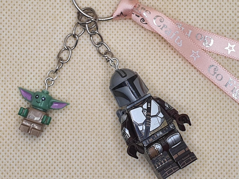 Puede incluir: Un llavero con dos figuras estilo Lego: El Mandaloriano y Grogu. La figura del Mandaloriano viste un traje plateado y marr&oacute;n. Grogu es verde con orejas grandes. El llavero tiene una cinta rosa con texto.