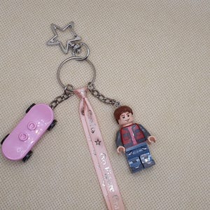 Könnte beinhalten: Ein Schlüsselanhänger mit einem rosa Skateboard, einer Lego-Figur und einem sternförmigen Anhänger. Der Schlüsselanhänger hat ein rosa Band mit Text und ist an einem silbernen Ring und einer Kette befestigt.