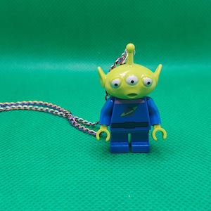 May include: A necklace featuring a green alien figure with three eyes, wearing a blue uniform. The alien is a Lego-style toy. The necklace chain is a multicolored braid, set against a green background.