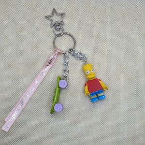 Może przedstawiać: Brelok do kluczy z srebrnym charms'em w kształcie gwiazdy, różową wstążką z tekstem, zielonym charms'em deskorolki z fioletowymi kołami oraz żółtym, czerwonym i niebieskim charms'em postaci Barta Simpsona.