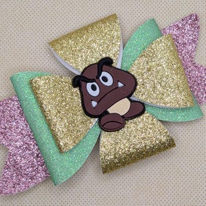 Puede incluir: Un lazo para el cabello hecho a mano con un personaje de Goomba. El lazo tiene capas de material dorado brillante, verde menta y rosa. El Goomba es marrón con una cara blanca y ojos negros. Accesorio para fans de videojuegos.