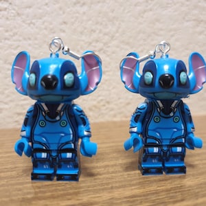 Puede incluir: Un par de pendientes azules con tem&aacute;tica de Stitch con un dise&ntilde;o de minifigura de Lego. La minifigura lleva un traje espacial azul y blanco con un casco azul. Los pendientes est&aacute;n hechos de pl&aacute;stico y tienen ganchos plateados.