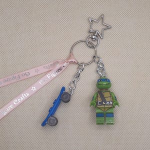 May include: A keyring featuring a green Teenage Mutant Ninja Turtle figure with a blue mask, a small blue plastic skateboard, and a light pink ribbon printed with the text Go Figure Crafts, all attached to a silver star-shaped metal clasp.