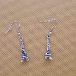 May include: A pair of silver Eiffel Tower earrings. Each earring has a detailed miniature replica of the Parisian landmark, suspended from a simple hook. The earrings are displayed against a neutral, beige background.
