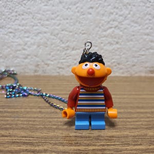 Könnte beinhalten: Eine Halskette mit einer Lego-ähnlichen Figur einer Zeichentrickfigur. Die Figur hat einen orangefarbenen Kopf, ein rotes Hemd, eine blaue Hose und schwarze Haare. Oben ist eine silberne Schlaufe befestigt. Eine mehrfarbige Kette liegt neben der Figur.