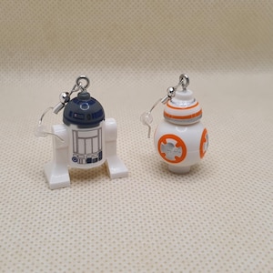 May include: A pair of earrings depicting Star Wars droids. One earring is a white and blue R2-D2 design, and the other is a white and orange BB-8 design. Both earrings have silver-coloured ear wires.