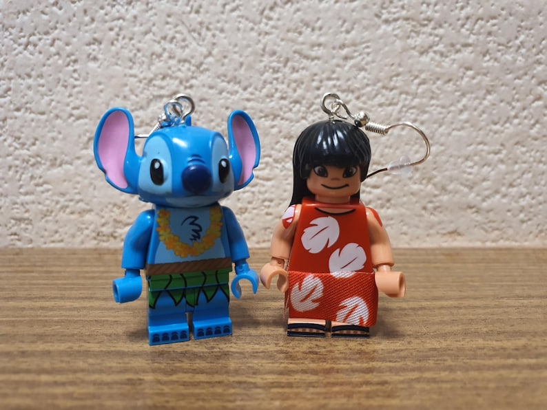 Może przedstawiać: Dwa kolczyki z postaciami w stylu Lego. Jeden jest niebieski z dużymi uszami i ż&oacute;łtym lei, drugi to dziewczyna w czerwonej sukience z białymi kwiatowymi wzorami. Oba mają srebrne haczyki.