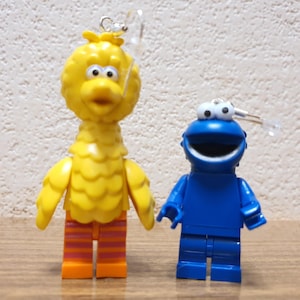 Könnte beinhalten: Ein Paar Ohrringe mit Sesamstraße-Charakteren, Big Bird und Cookie Monster. Big Bird ist gelb mit orange-rosa gestreiften Hosen. Cookie Monster ist blau mit einem weit geöffneten Mund und weißen Augen.