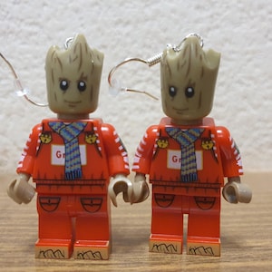 May include: A pair of orange and white Lego-style earrings featuring a character with a brown face and a green scarf. The character is wearing a red and white striped shirt and has a green scarf around its neck. The earrings are attached to silver hooks.