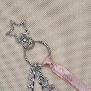 Puede incluir: Llavero plateado con un cierre en forma de estrella, una cadena y una cinta rosa. La cinta tiene texto impreso. El llavero est&aacute; dise&ntilde;ado para guardar llaves u otros objetos peque&ntilde;os. El fondo es de color beige claro.