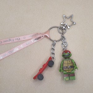 Op de afbeelding: Een sleutelhanger met een rode skateboard-bedel en een groene actiefiguur van een schildpad met een rood masker. De sleutelhanger bevat ook een zilveren sterbedel en een roze lint met de tekst "Go Figure Crafts".