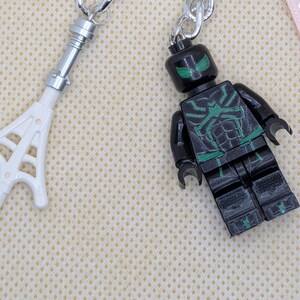 May include: A black and green plastic minifigure keychain featuring a spider-like chest emblem and green eyes, paired with a white plastic web accessory attached to a silver metal ring, showcasing detailed molded features and articulated limbs.
