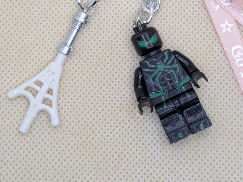May include: A black and green plastic minifigure keychain featuring a spider-like chest emblem and green eyes, paired with a white plastic web accessory attached to a silver metal ring, showcasing detailed molded features and articulated limbs.