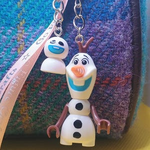 Puede incluir: Dos llaveros con el personaje Olaf de la pel&iacute;cula Frozen. Uno muestra la cabeza de Olaf con una cara sonriente, y el otro muestra el cuerpo completo de Olaf. Los llaveros est&aacute;n unidos a una cadena plateada, sobre un fondo azul y morado.