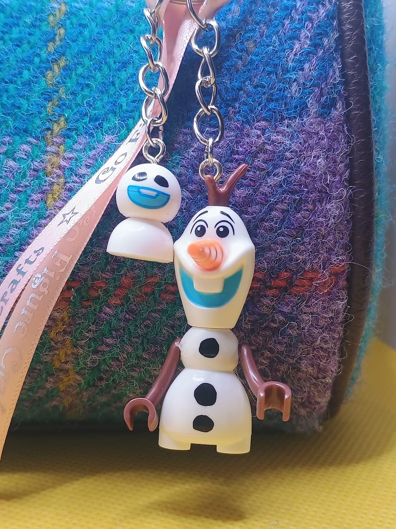 Puede incluir: Dos llaveros con el personaje Olaf de la pel&iacute;cula Frozen. Uno muestra la cabeza de Olaf con una cara sonriente, y el otro muestra el cuerpo completo de Olaf. Los llaveros est&aacute;n unidos a una cadena plateada, sobre un fondo azul y morado.