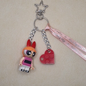 May include: A keychain featuring a cartoon character figure with red hair and a pink and black outfit, a red heart-shaped charm, and a silver star-shaped clasp. The keychain includes a pink ribbon with text.