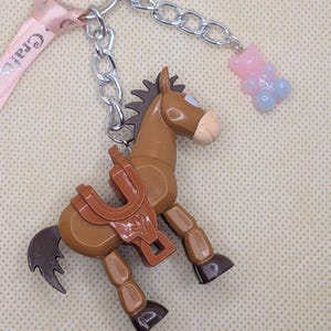 May include: A brown horse-shaped keychain with a saddle, attached to a silver chain and a pink ribbon with the text "Crafts". A small pink and blue gummy bear charm hangs from the chain.