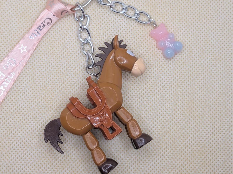 May include: A brown horse-shaped keychain with a saddle, attached to a silver chain and a pink ribbon with the text "Crafts". A small pink and blue gummy bear charm hangs from the chain.
