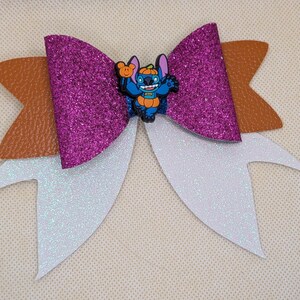 Può includere: Un fiocco decorativo con uno strato centrale di glitter viola, uno strato marrone testurizzato e uno strato di glitter bianco. Un personaggio blu con un cappello a forma di zucca arancione e una piccola zucca è posto al centro del fiocco.