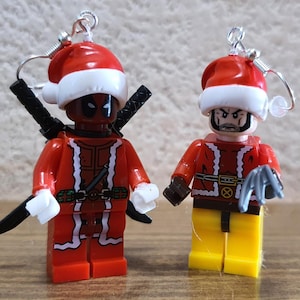 May include: Two Christmas-themed Lego minifigures, one dressed as Deadpool and the other as Wolverine, both wearing Santa hats and red and white suits. The figures are designed as earrings.