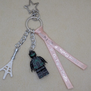 May include: A keychain featuring a silver star-shaped clasp, a metal ring, a white plastic web accessory on a chain, a black and green minifigure on a chain, and a light pink ribbon printed with the text Go Figure Crafts and small stars.
