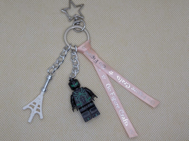 May include: A keychain featuring a silver star-shaped clasp, a metal ring, a white plastic web accessory on a chain, a black and green minifigure on a chain, and a light pink ribbon printed with the text Go Figure Crafts and small stars.