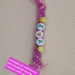 May include: A personalized keychain with the name "EVA" spelled out in white letter beads. The keychain features a braided purple and pink cord, with yellow beads and a silver keyring. A pink tag reads "Example of additional personalised charm. Colours will vary."