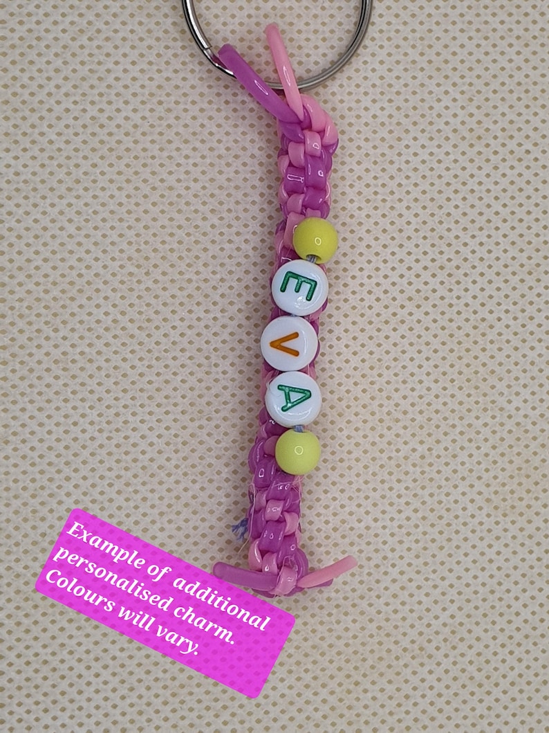 May include: A personalized keychain with the name "EVA" spelled out in white letter beads. The keychain features a braided purple and pink cord, with yellow beads and a silver keyring. A pink tag reads "Example of additional personalised charm. Colours will vary."