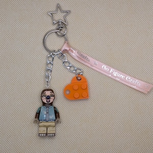 Puede incluir: Un llavero plateado con una figura de perezoso, un dije en forma de corazón naranja y un cierre en forma de estrella. La figura del perezoso lleva una camisa azul y pantalones color caqui. Una cinta rosa con "Go Figure Crafts" está adjunta.