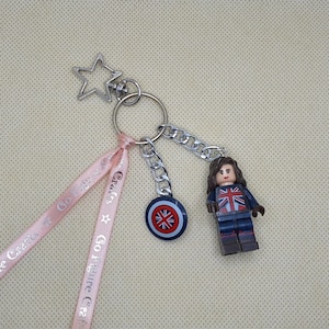 May include: A silver-tone keychain with a star-shaped clasp, a chain, and a small shield charm. Attached is a Lego figure wearing a Union Jack design. Pink ribbons with the text "Go Figure Crafts" are also attached.