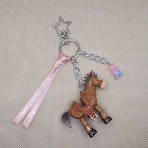 May include: A brown horse keychain with a saddle, a star-shaped charm, and a pink ribbon with the text "Figure Crafts". A small chain connects the horse to a gummy bear charm.