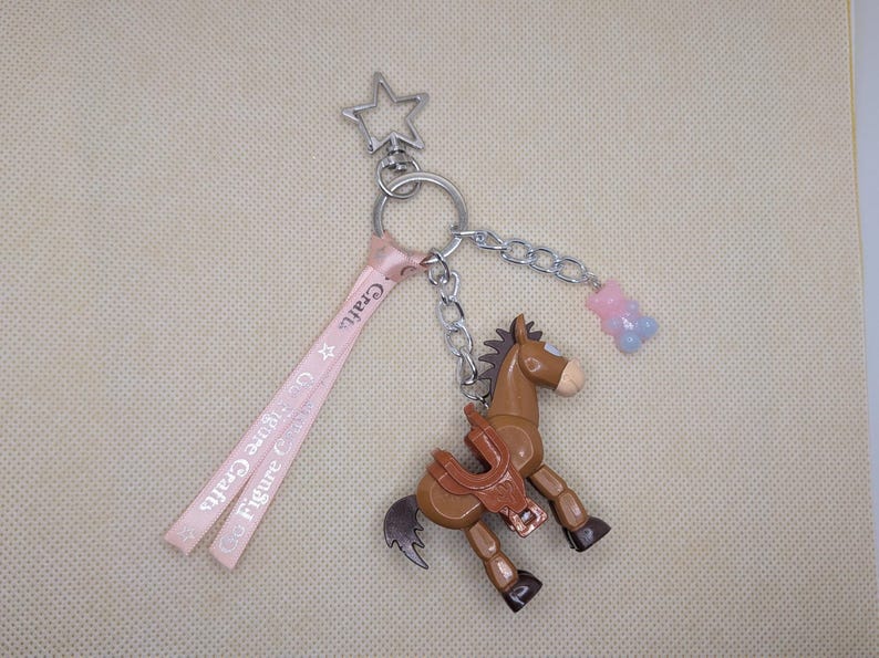 May include: A brown horse keychain with a saddle, a star-shaped charm, and a pink ribbon with the text "Figure Crafts". A small chain connects the horse to a gummy bear charm.