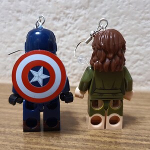 May include: A pair of earrings featuring Lego minifigures. One earring features Captain America with his signature shield and the other earring features a female minifigure with brown hair and a green jacket.