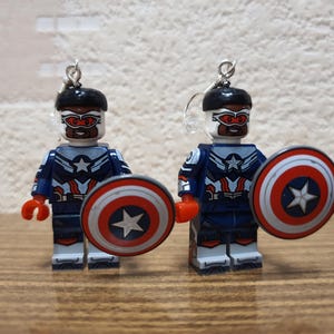 May include: A pair of earrings featuring miniature Captain America figures made from toy blocks. Each figure is wearing a blue and white uniform, a black helmet, and holding a shield with a red, white, and blue design. The earrings have silver-coloured hooks.