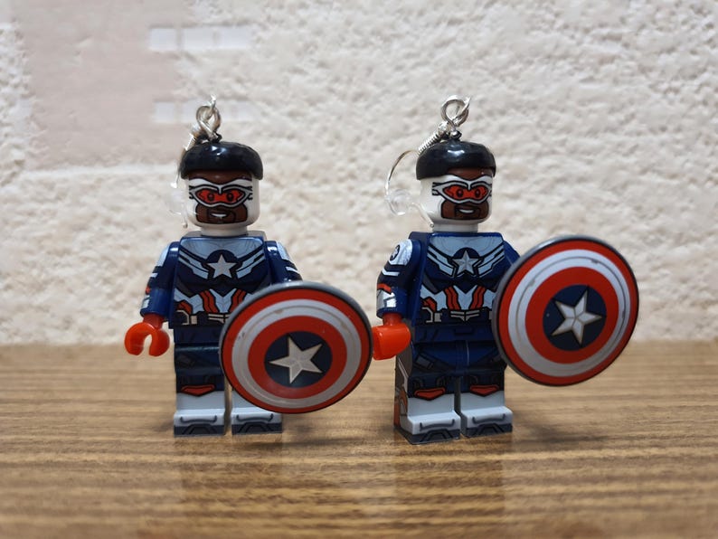 May include: A pair of earrings featuring miniature Captain America figures made from toy blocks. Each figure is wearing a blue and white uniform, a black helmet, and holding a shield with a red, white, and blue design. The earrings have silver-coloured hooks.