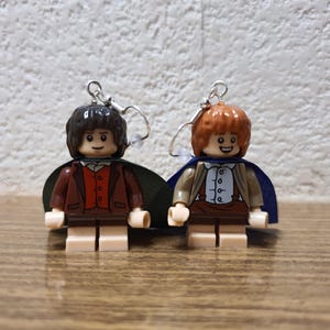 Puede incluir: Un par de pendientes con minifiguras Lego de personajes de El Señor de los Anillos. La minifigura de la izquierda lleva una chaqueta marrón y una capa verde, mientras que la minifigura de la derecha lleva una chaqueta marrón y una capa azul.