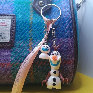 Puede incluir: Un llavero plateado con dos colgantes: un mu&ntilde;eco de nieve sonriente y una peque&ntilde;a figura blanca con una cara azul. El llavero est&aacute; sujeto a un bolso de tweed azul, morado y verde. Tambi&eacute;n hay una cinta rosa con texto.