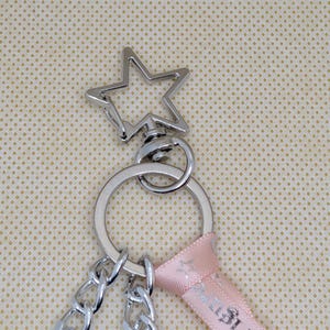 May include: A silver-toned metal keychain featuring a star-shaped lobster clasp attached to a circular split ring. The hardware is connected to a light pink fabric ribbon and two silver-toned metal chains that hang downward from the ring.