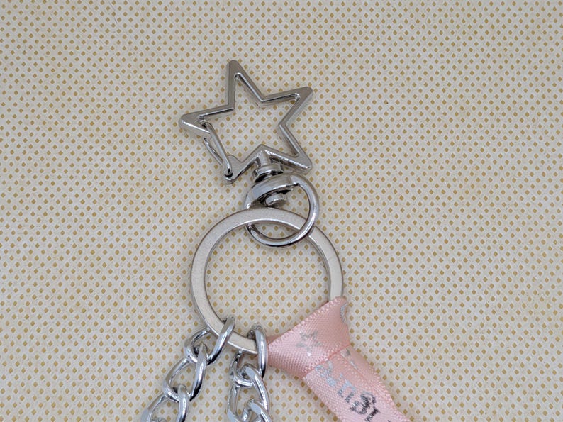 May include: A silver-toned metal keychain featuring a star-shaped lobster clasp attached to a circular split ring. The hardware is connected to a light pink fabric ribbon and two silver-toned metal chains that hang downward from the ring.