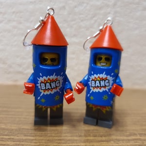 Puede incluir: Un par de pendientes de minifigura Lego azul y rojo. Las minifiguras están vestidas como cohetes con la palabra "BANG" en el pecho.