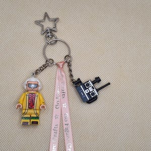 Könnte beinhalten: Ein Schlüsselanhänger mit einem silbernen Stern, einer Miniaturfigur in einer gelben Jacke und einer schwarzen Kamera. Die Figur hat einen weißen Kopf und trägt eine Brille. Ein rosa Band mit Text ist am Schlüsselanhänger befestigt.