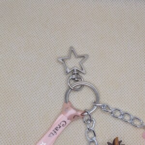 May include: Silver-toned keychain with a star-shaped top. A pink ribbon with the word "Crafts" is attached, along with a chain and various charms, including a bear and a small brown object. The keychain is designed for attaching to keys or bags.