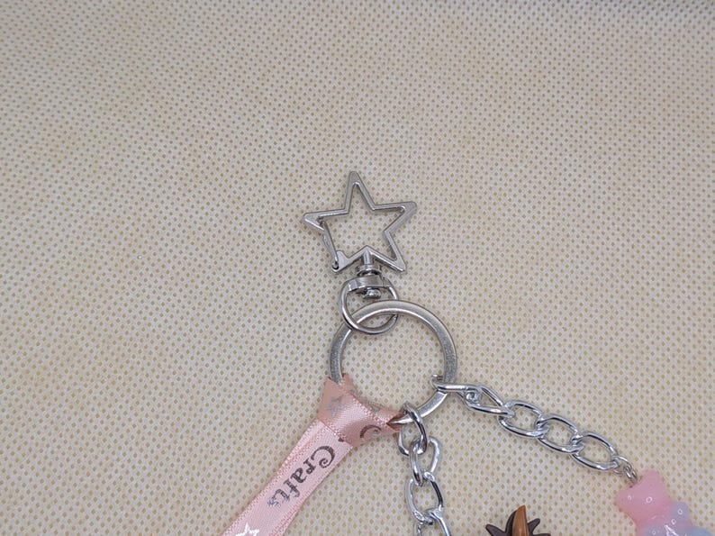 May include: Silver-toned keychain with a star-shaped top. A pink ribbon with the word "Crafts" is attached, along with a chain and various charms, including a bear and a small brown object. The keychain is designed for attaching to keys or bags.