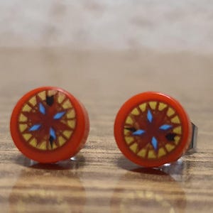 Puede incluir: Un par de pendientes de botón rojos con un diseño de estrella azul y dorada.