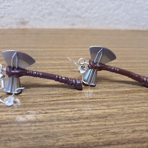 May include: A pair of silver axe earrings with brown handles. The earrings are on a wooden surface.