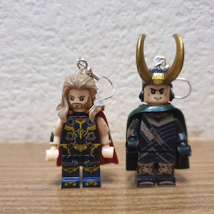 Pode incluir: Um par de brincos com minifiguras Lego de Thor e Loki. Thor está usando sua capa vermelha e capacete característicos, enquanto Loki está usando sua capa verde e chifres.