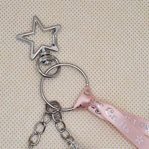 Puede incluir: Llavero plateado con una parte superior en forma de estrella y una cadena. Una cinta rosa con el texto "Crafts" y dise&ntilde;os de estrellas est&aacute; unida al llavero. El fondo es una tela beige claro.