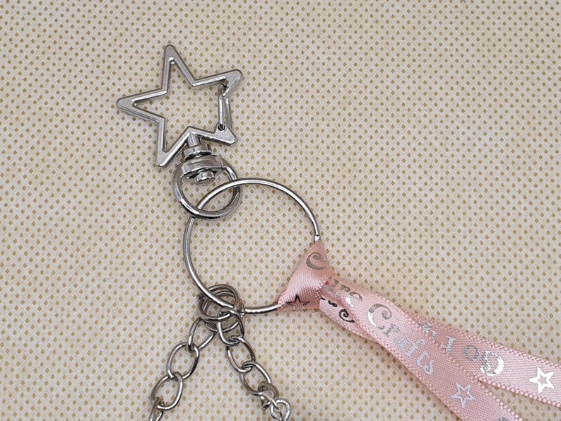 Puede incluir: Llavero plateado con una parte superior en forma de estrella y una cadena. Una cinta rosa con el texto "Crafts" y dise&ntilde;os de estrellas est&aacute; unida al llavero. El fondo es una tela beige claro.